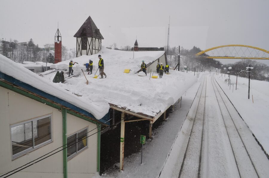 Cuadrilla despejando con palas la cubierta nevada de una estación, con el canto del manto a la vista en el alero